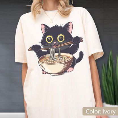May include: Ivory-colored t-shirt featuring a cartoon black cat eating ramen noodles with chopsticks. The cat has large yellow eyes and pink paws. The ramen bowl is beige with brown noodles. The text "Color: Ivory" is at the bottom.