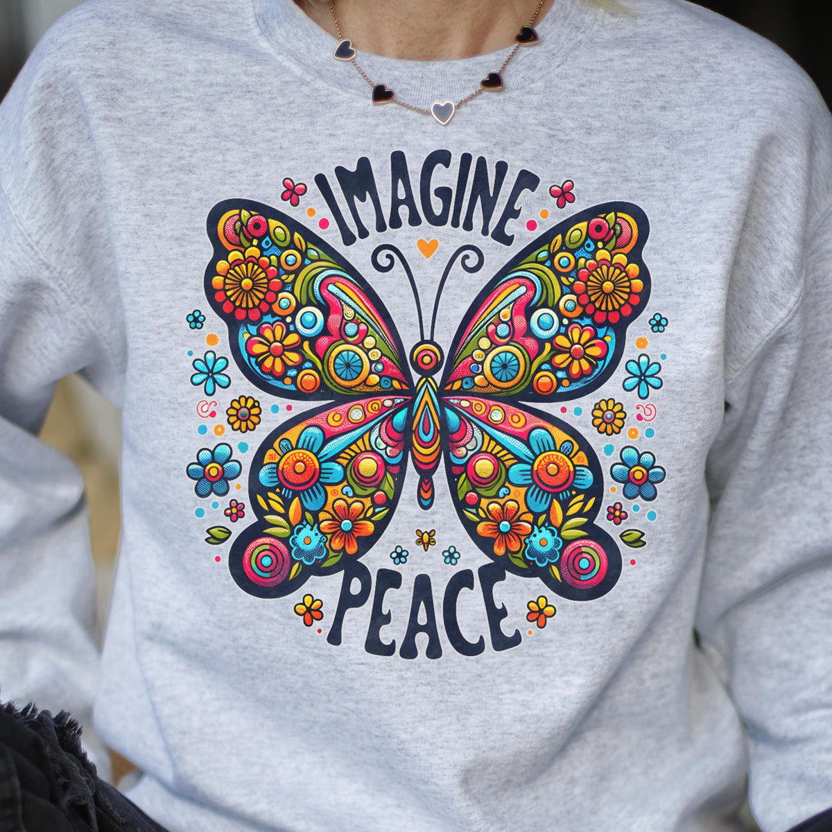 May include: A light gray sweatshirt featuring a vibrant butterfly design with the words "IMAGINE PEACE". The butterfly is adorned with colorful floral patterns in shades of blue, orange, yellow, and pink. The sweatshirt has a crew neck.