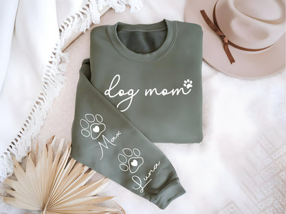 May include: Olive green sweatshirt with the words "dog mom" and a paw print graphic. The sleeve features paw prints with the names "Max" and "Luna". The sweatshirt is folded on a white surface with a hat in the background.