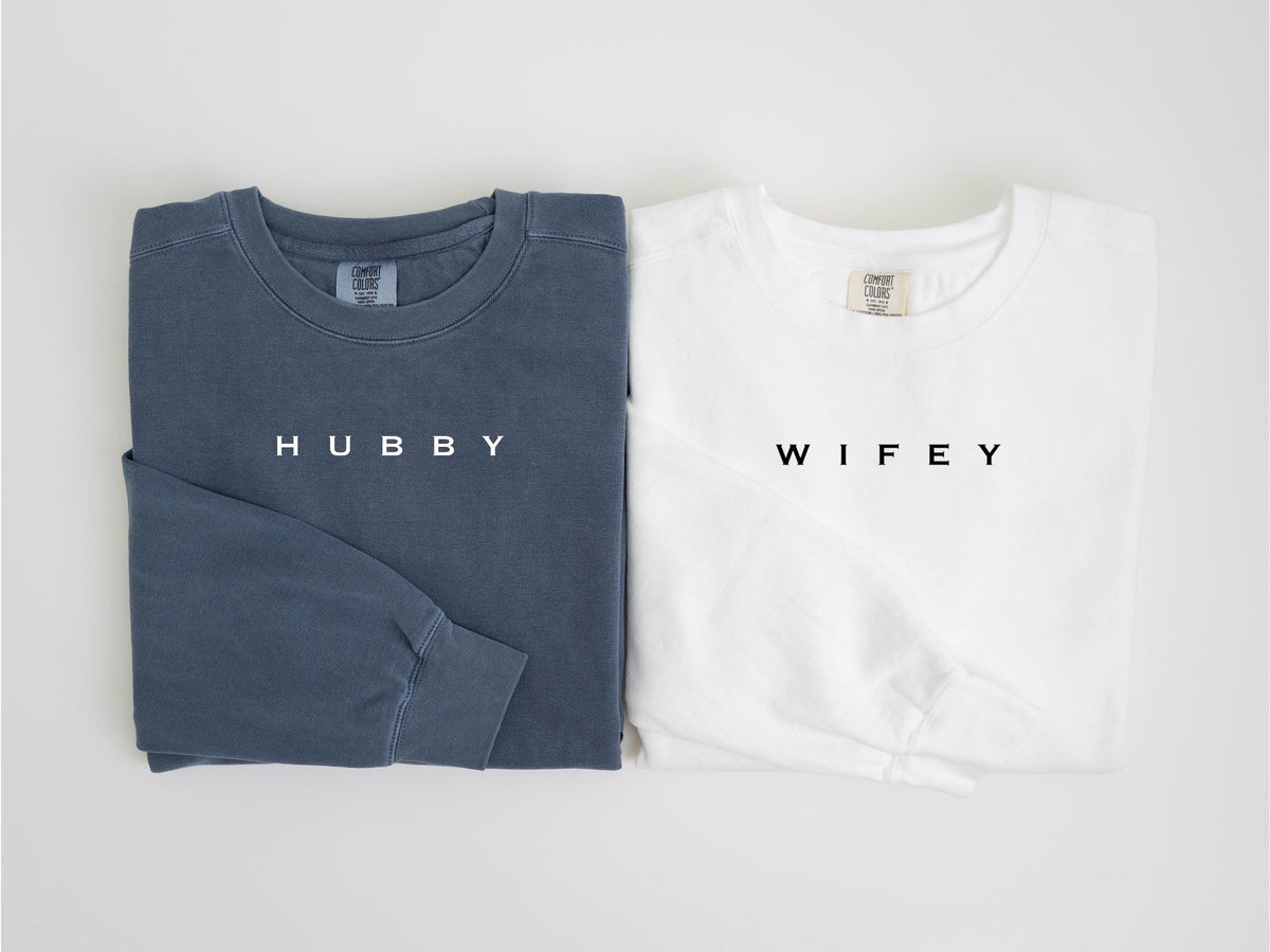 May include: Two long-sleeved shirts, one in a muted blue-gray and the other in white. The blue-gray shirt has the word "HUBBY" printed in white, while the white shirt has "WIFEY" printed in black. Both shirts are folded.