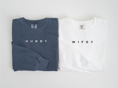 May include: Two long-sleeved shirts, one in a muted blue-gray and the other in white. The blue-gray shirt has the word "HUBBY" printed in white, while the white shirt has "WIFEY" printed in black. Both shirts are folded.