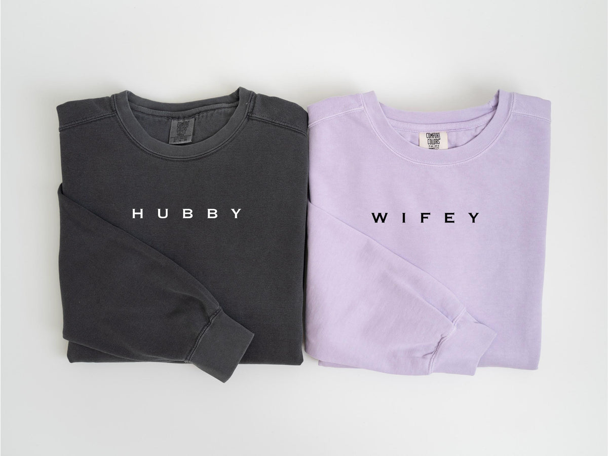 May include: Two long-sleeved shirts, one charcoal gray and one lavender, are displayed. The gray shirt has the word "HUBBY" printed in white, while the lavender shirt has "WIFEY" printed in black. Both shirts are folded neatly.