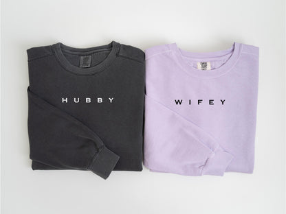 May include: Two long-sleeved shirts, one charcoal gray and one lavender, are displayed. The gray shirt has the word "HUBBY" printed in white, while the lavender shirt has "WIFEY" printed in black. Both shirts are folded neatly.