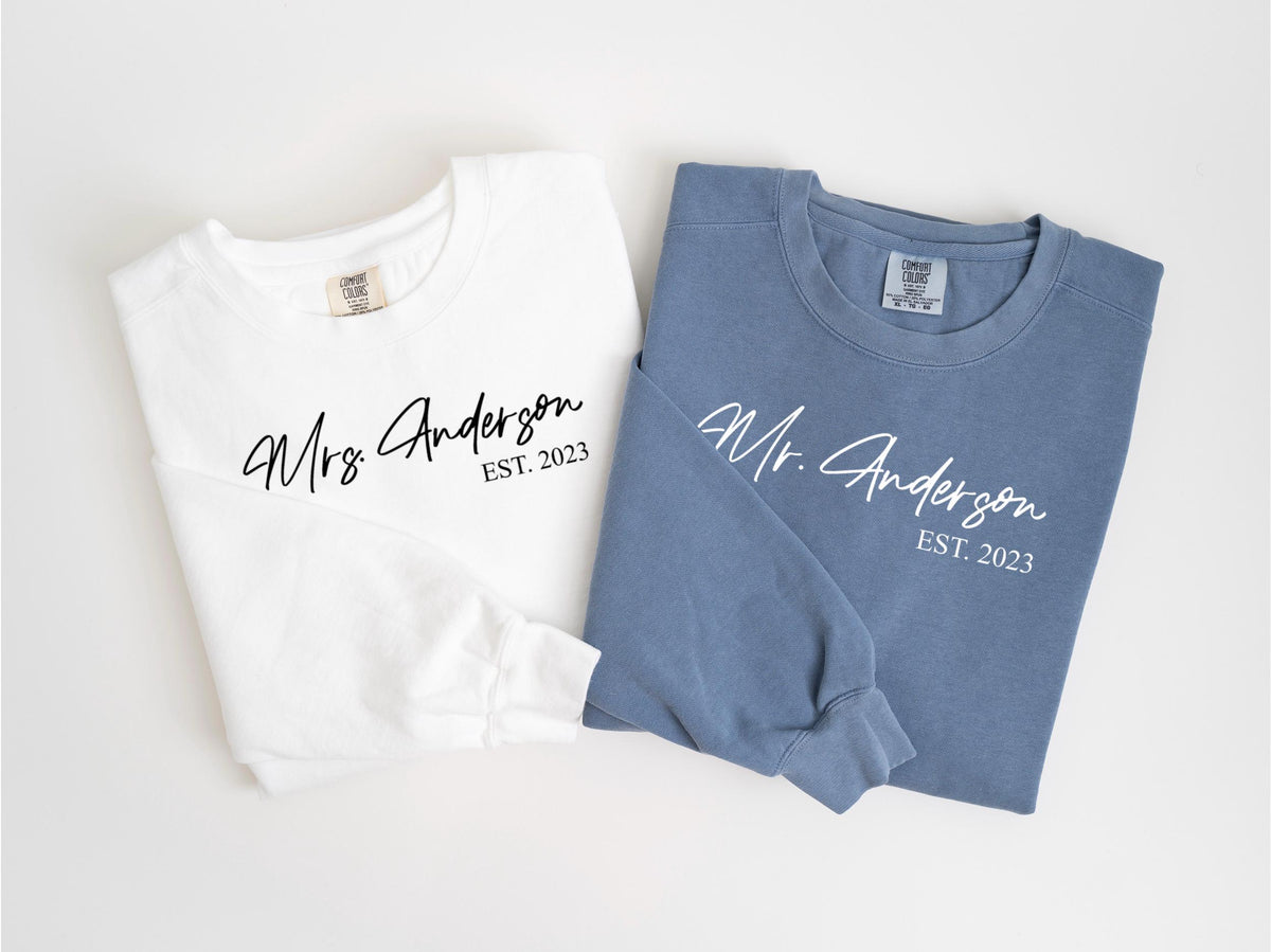 May include: Two long-sleeved sweatshirts, one white and one blue, with the names "Mrs. Anderson" and "Mr. Anderson" printed in cursive, along with "EST. 2023". The shirts are likely for a couple.