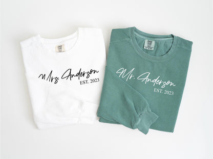 May include: Two matching sweatshirts, one white and one green, with the names "Mrs. Anderson" and "Mr. Anderson" printed in cursive, along with "EST. 2023". The white sweatshirt is on the left, and the green one is on the right.