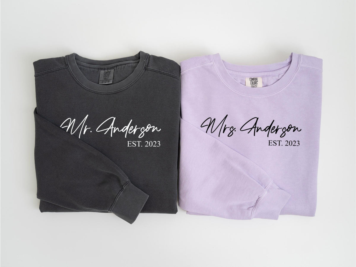 May include: Two long-sleeved shirts, one charcoal gray and one lavender, are displayed. The gray shirt reads "Mr. Anderson EST. 2023" in white script. The lavender shirt reads "Mrs. Anderson EST. 2023" in black script.