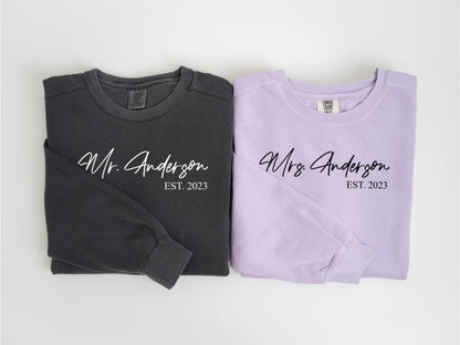 May include: Two long-sleeved shirts, one charcoal gray and one lavender, are displayed. The gray shirt reads "Mr. Anderson EST. 2023" in white script. The lavender shirt reads "Mrs. Anderson EST. 2023" in black script.