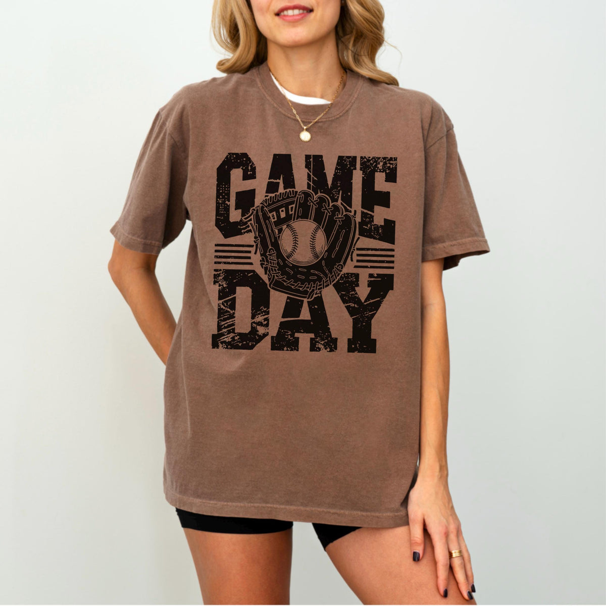 May include: Brown t-shirt with the words "GAME DAY" in a distressed black font. A baseball glove and ball graphic is centered between the words. The shirt is a solid color and has short sleeves.
