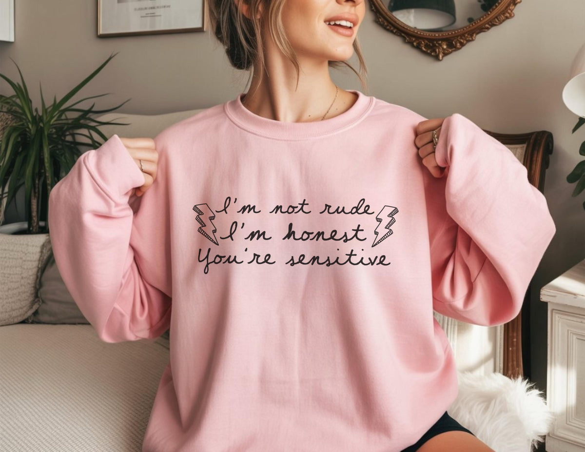 May include: A light pink sweatshirt with the text "I'm not rude, I'm honest, You're sensitive" in black script. The text is framed by two lightning bolt graphics. The sweatshirt has a crew neck and long sleeves.