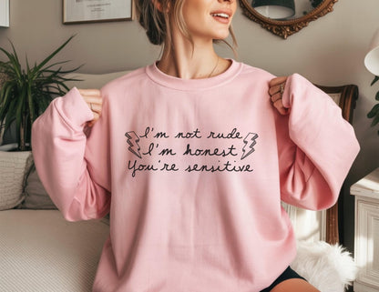 May include: A light pink sweatshirt with the text "I'm not rude, I'm honest, You're sensitive" in black script. The text is framed by two lightning bolt graphics. The sweatshirt has a crew neck and long sleeves.
