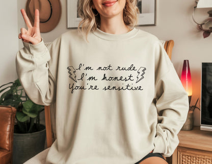 May include: A beige sweatshirt with the text "I'm not rude, I'm honest, You're sensitive" in black script. The design includes lightning bolt graphics. The sweatshirt has long sleeves and a crew neck.