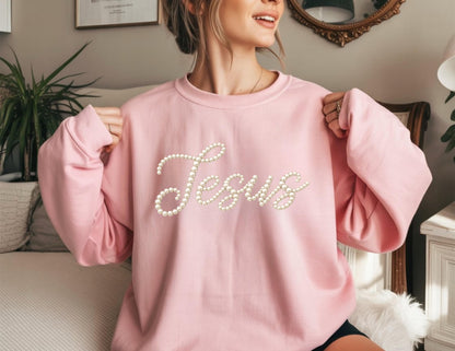 May include: A light pink sweatshirt with the word "Jesus" spelled out in white pearl-like beads. The sweatshirt has long sleeves and a crew neck. The overall style is casual and comfortable.
