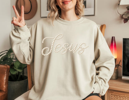 May include: A light beige sweatshirt with the word "Jesus" spelled out in white pearls. The long-sleeved top is a neutral color and has a crew neck. The sweatshirt is a casual style.
