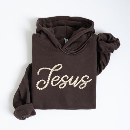 May include: A dark brown hoodie with the word "Jesus" spelled out in off-white faux pearls. The hoodie has a drawstring hood and long sleeves. The garment is folded neatly on a white surface.
