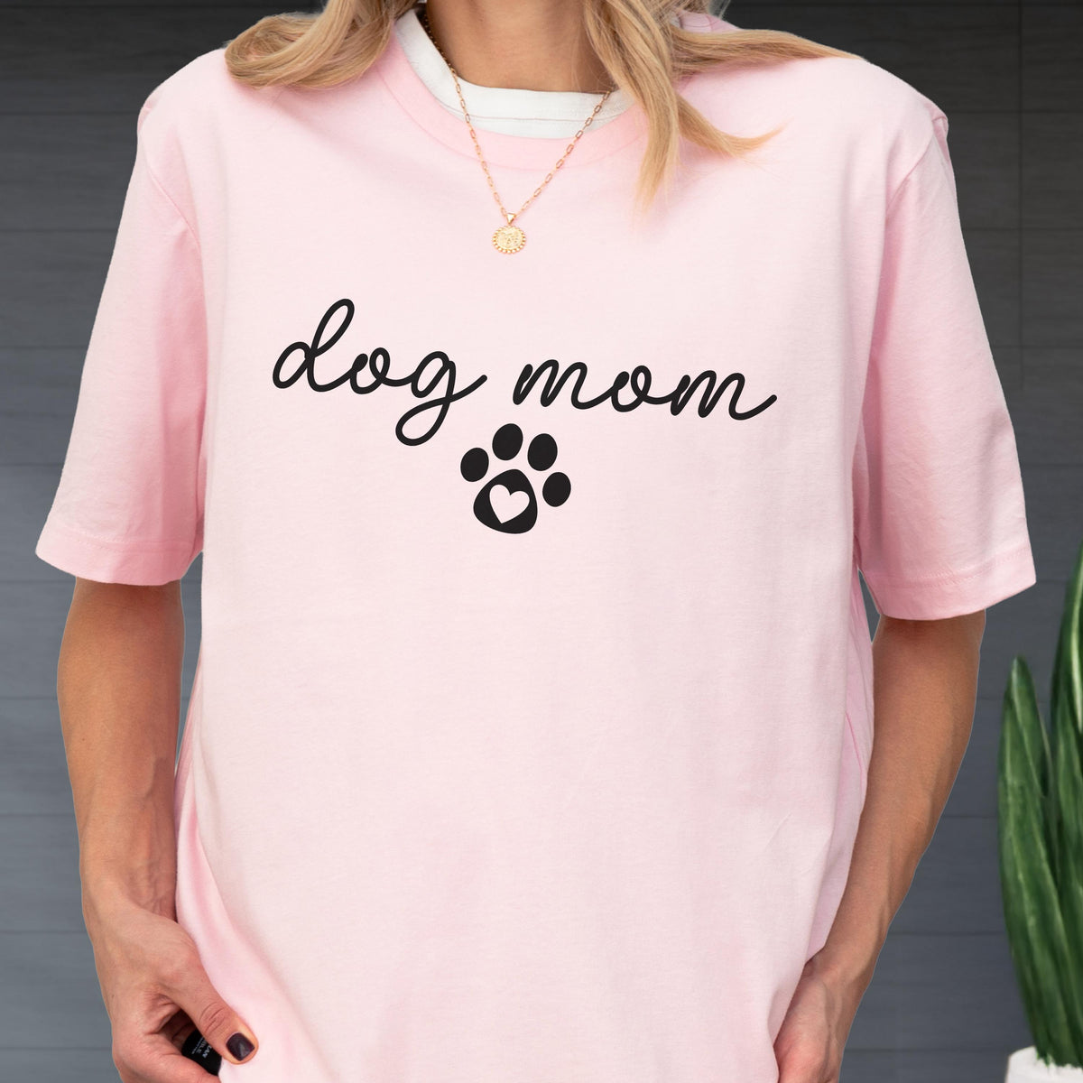 May include: A light pink t-shirt with the words "dog mom" in a cursive font, and a paw print with a heart in the center. The shirt has short sleeves and a crew neck.