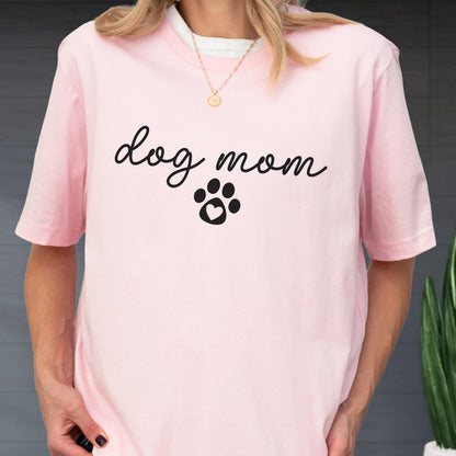 May include: A light pink t-shirt with the words "dog mom" in a cursive font, and a paw print with a heart in the center. The shirt has short sleeves and a crew neck.