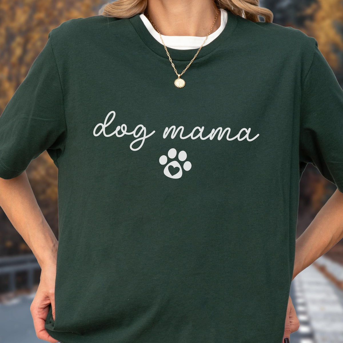 May include: Dark green t-shirt with the words "dog mama" in cursive script and a white paw print graphic. The shirt has a white crew neck collar and is worn with a gold necklace.