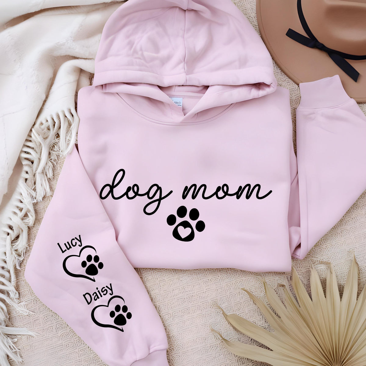 May include: A light pink hooded sweatshirt with the words "dog mom" in black script, a paw print with a heart, and the names "Lucy" and "Daisy" with paw prints inside hearts on the sleeve. The sweatshirt is folded on a cream-colored blanket.