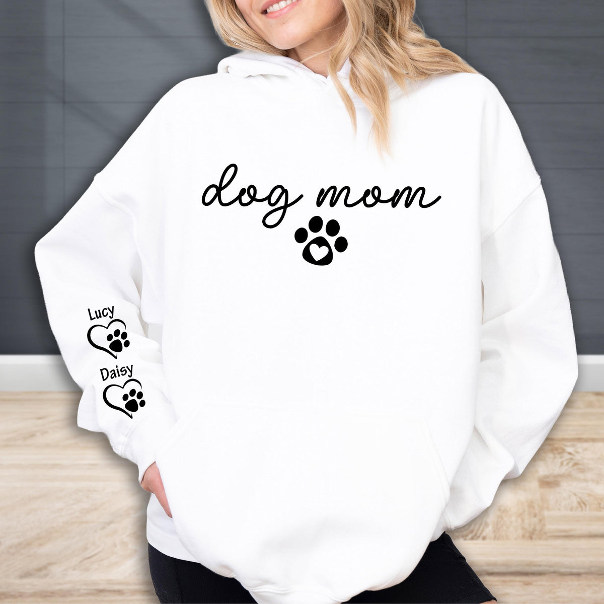 May include: White hoodie with the words "dog mom" in a script font, a paw print with a heart, and two paw print hearts with the names "Lucy" and "Daisy" on the sleeve. The hoodie is a casual style.