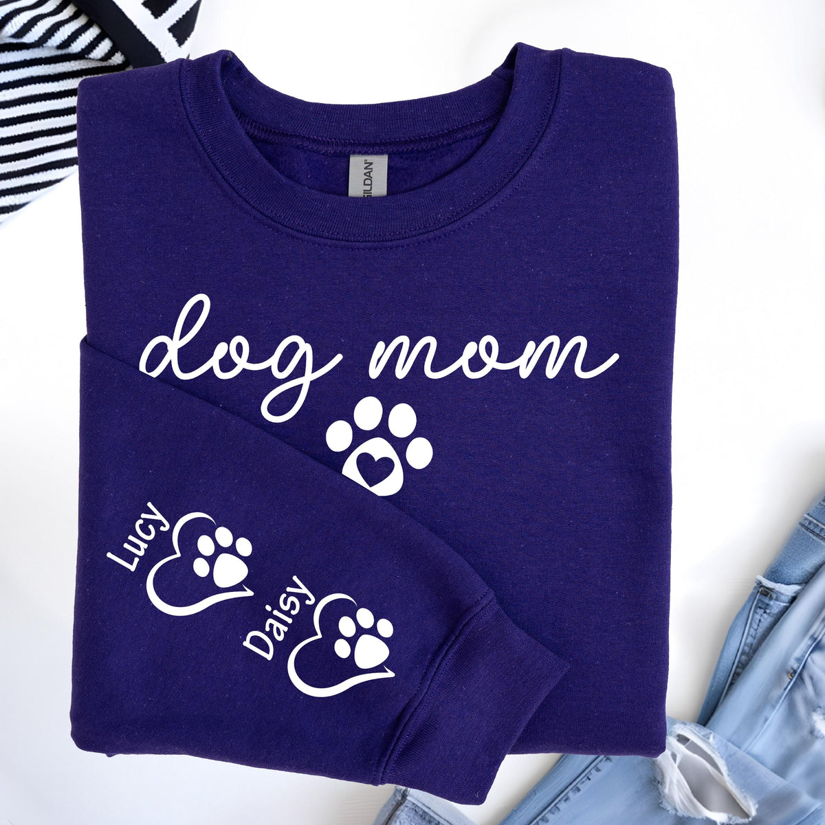 May include: A dark purple sweatshirt with the words "dog mom" in white script, along with a paw print with a heart. The sleeve features the names "Lucy" and "Daisy" with paw prints inside hearts. The sweatshirt is made of a soft material.