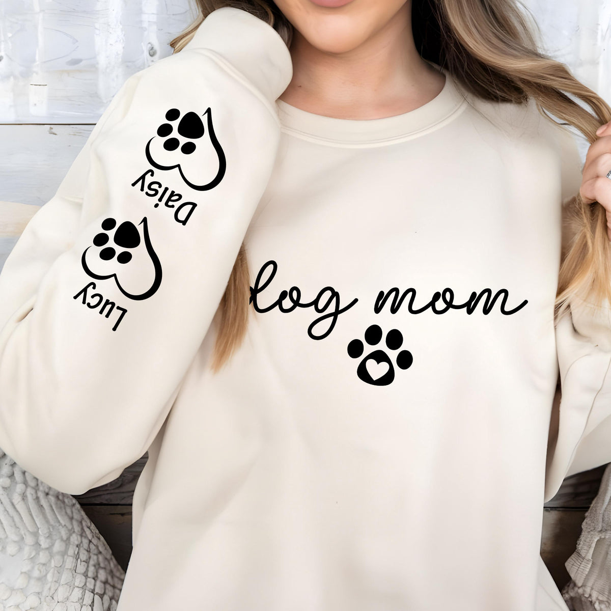 Model wearing a sand-colored crewneck sweatshirt with 'Dog Mom' and a paw print on the chest. The sleeve features personalized pet names 'Daisy' and 'Lucy' next to heart-shaped paw prints. Perfect custom gift for dog lovers.