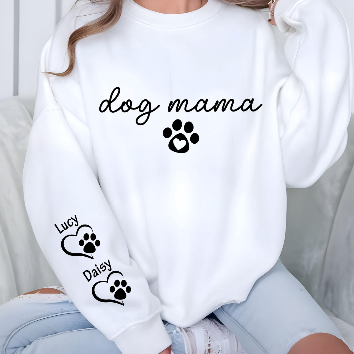 May include: White sweatshirt with the text "dog mama" in a script font, a paw print with a heart, and two paw print hearts with the names "Lucy" and "Daisy" on the sleeve. The design is in black.