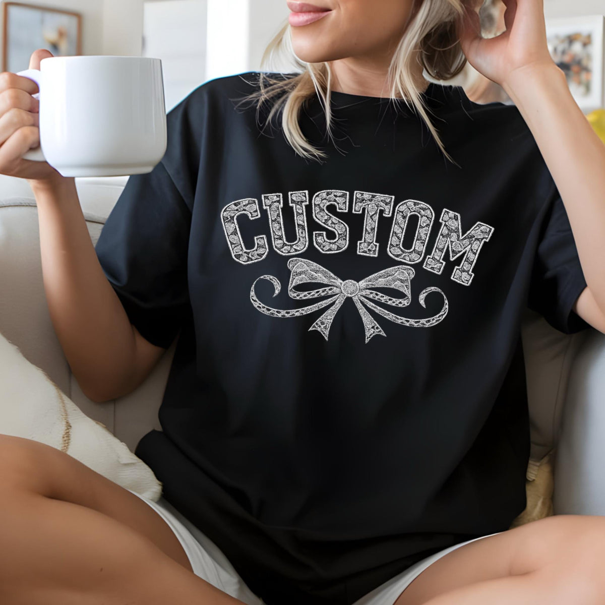 May include: Black t-shirt with the word "CUSTOM" in a white, decorative font above a detailed bow design. The shirt is worn by a person holding a white mug.