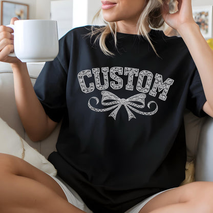 May include: Black t-shirt with the word "CUSTOM" in a white, decorative font above a detailed bow design. The shirt is worn by a person holding a white mug.
