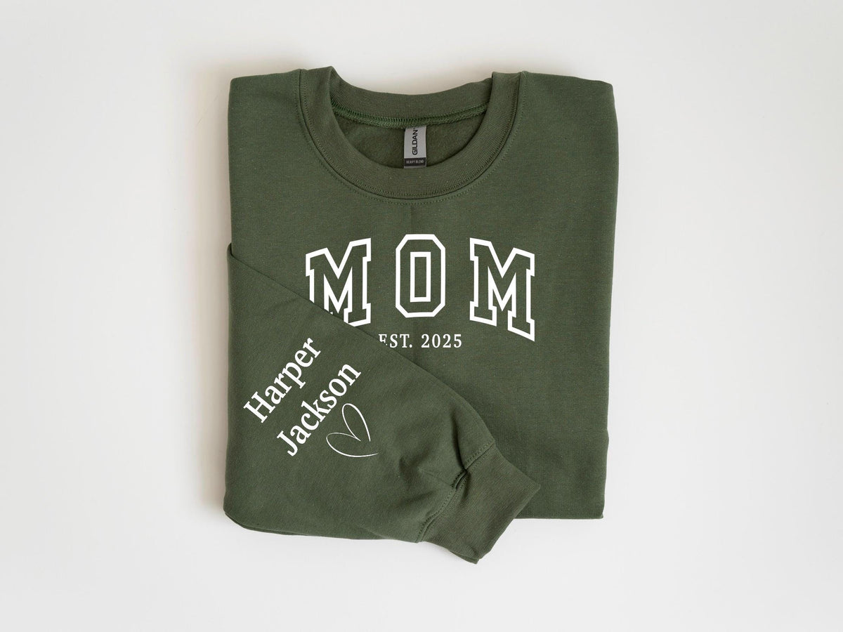 May include: An olive green sweatshirt with the word "MOM" in white block letters, with "EST. 2025" below. The left sleeve has the name "Harper Jackson" and a heart symbol printed in white. The sweatshirt is folded.