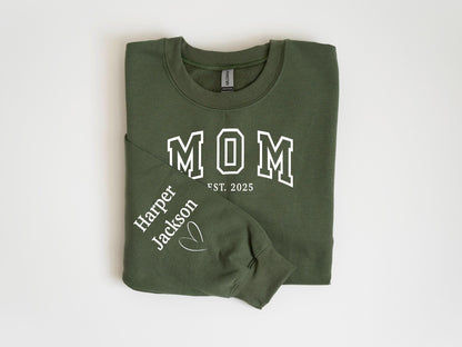 May include: An olive green sweatshirt with the word "MOM" in white block letters, with "EST. 2025" below. The left sleeve has the name "Harper Jackson" and a heart symbol printed in white. The sweatshirt is folded.