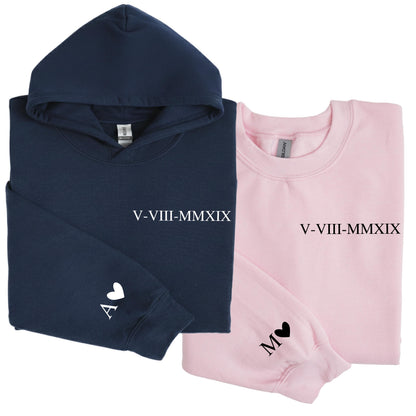May include: A navy blue hooded sweatshirt and a light pink crewneck sweatshirt. The navy hoodie features the white text V-VIII-MMXIX and a white letter A with a heart. The pink sweatshirt features black text V-VIII-MMXIX and a black letter M with a heart.