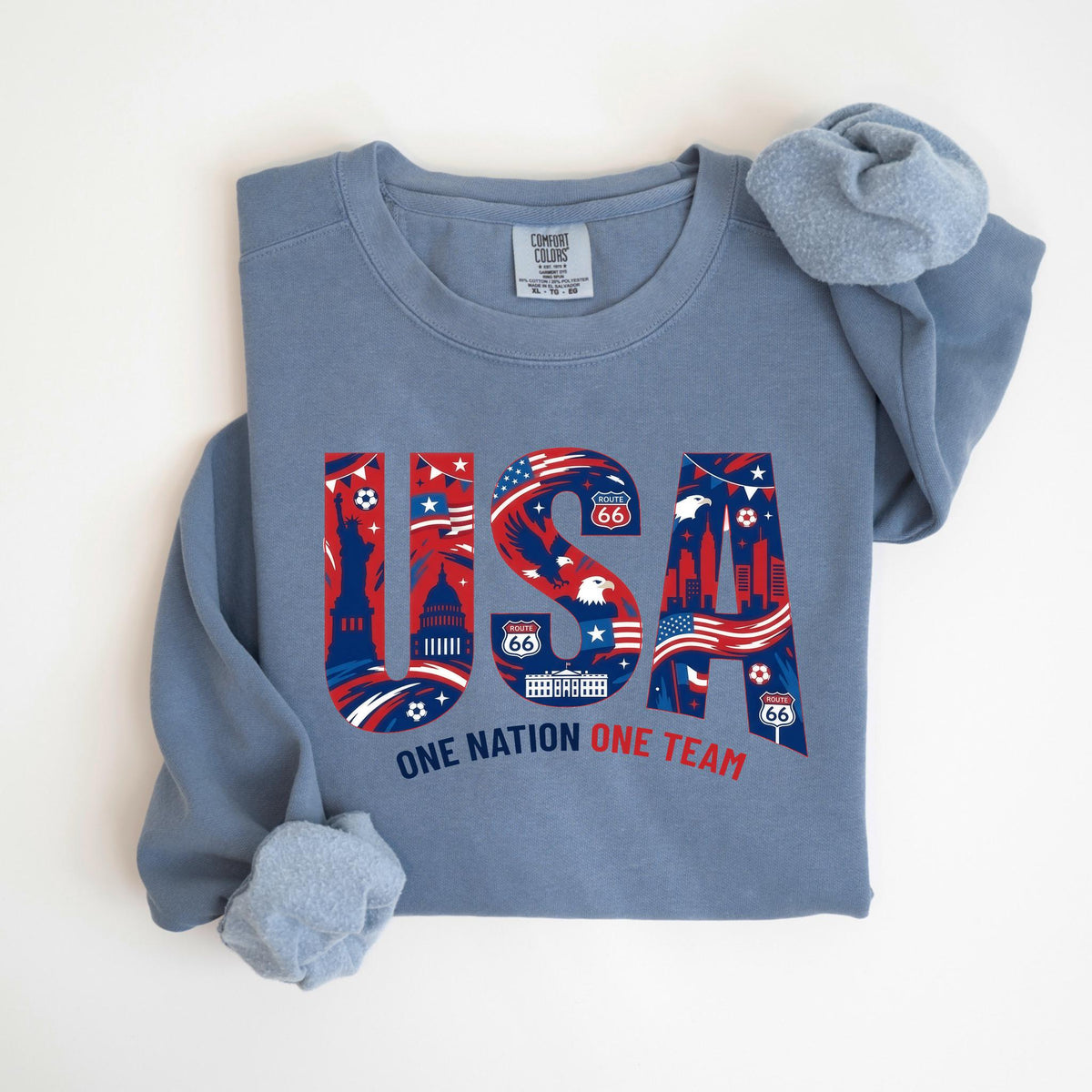 May include: A blue crew neck sweatshirt with the word "USA" in red, white, and blue, featuring iconic American imagery. The text "ONE NATION ONE TEAM" is below the word "USA".