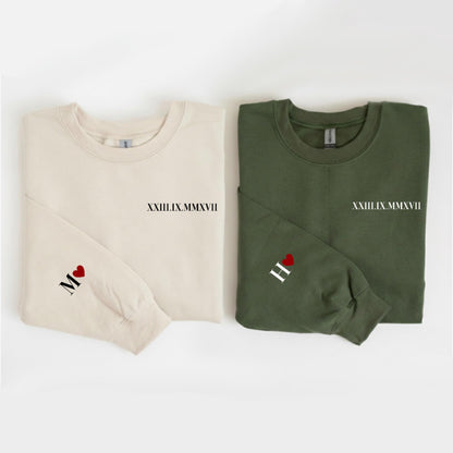 May include: Two long-sleeved sweatshirts, one cream and one olive green. Each has a date in Roman numerals and a single initial with a heart on the sleeve. The cream sweatshirt has the initial "M" and the olive green has the initial "H".