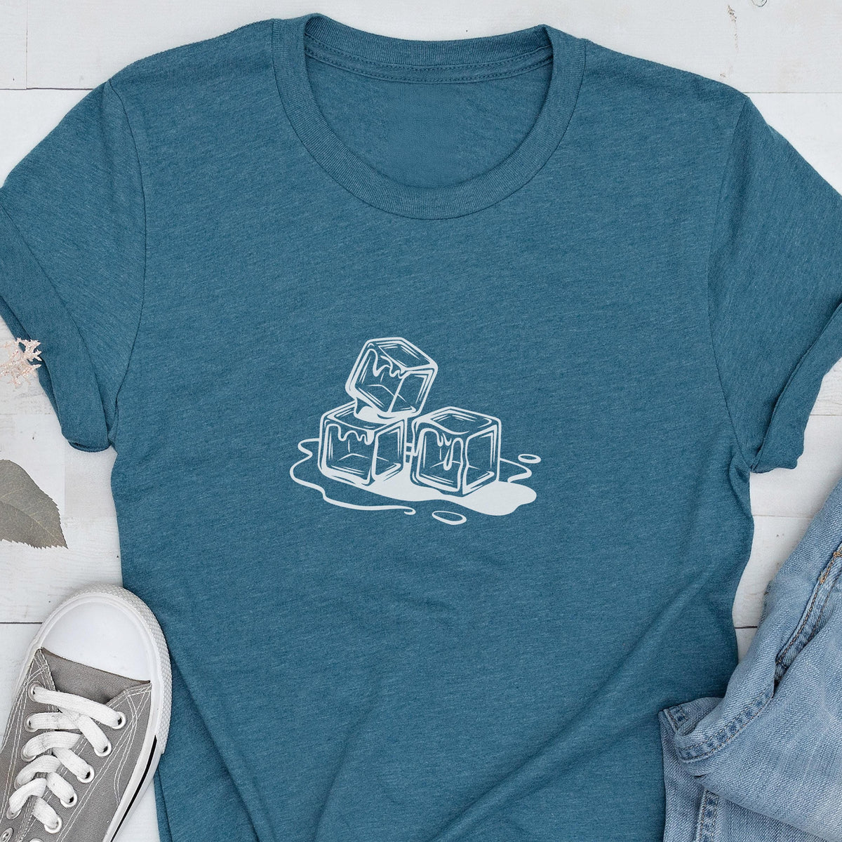 May include: Teal t-shirt featuring a white graphic of three ice cubes melting. The design is a simple, line-art style. The shirt has a crew neck and short sleeves. A pair of gray sneakers and blue jeans are visible in the image.