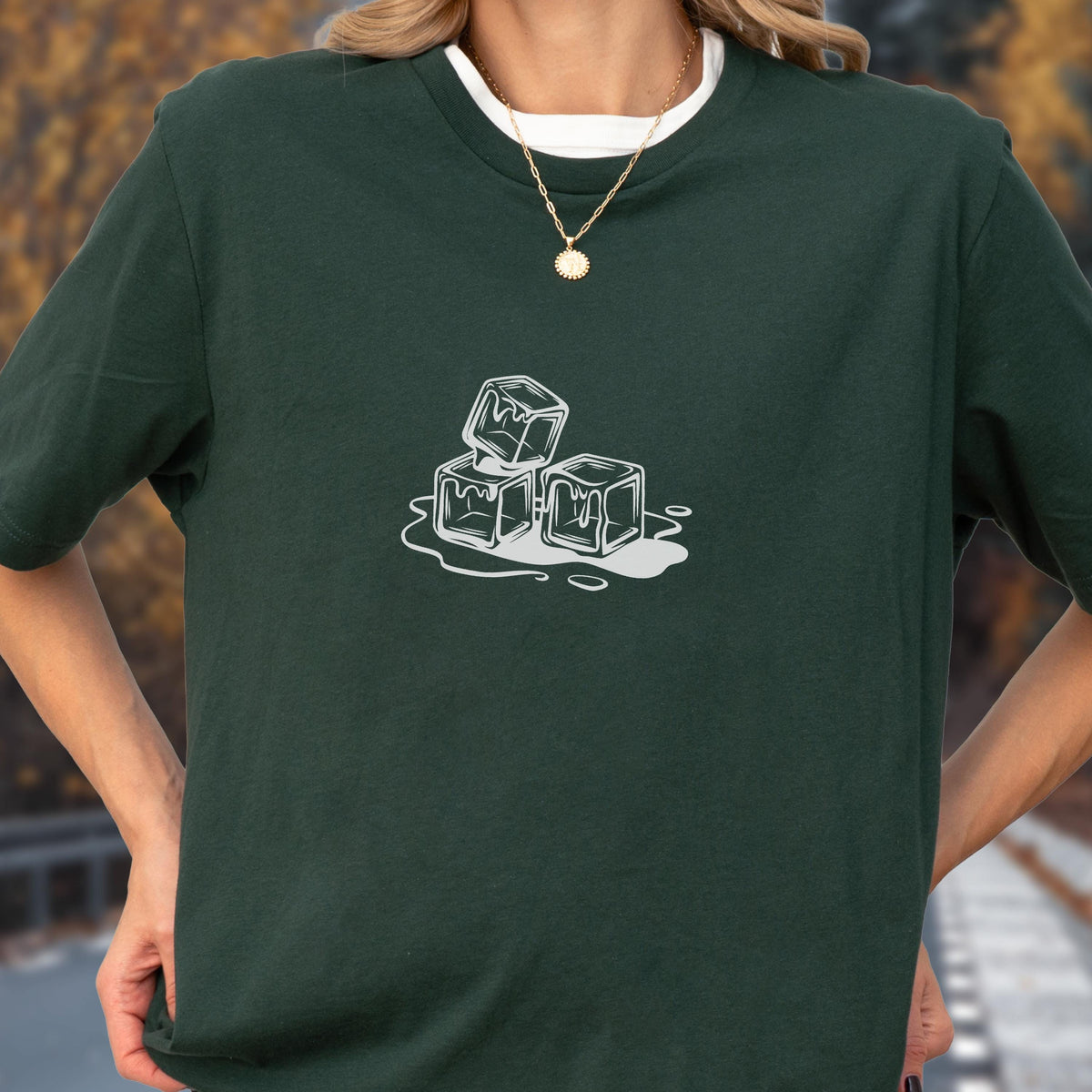 May include: Dark green t-shirt featuring a white graphic of three ice cubes with water droplets. The shirt has a crew neck and short sleeves. A gold necklace with a pendant is visible.