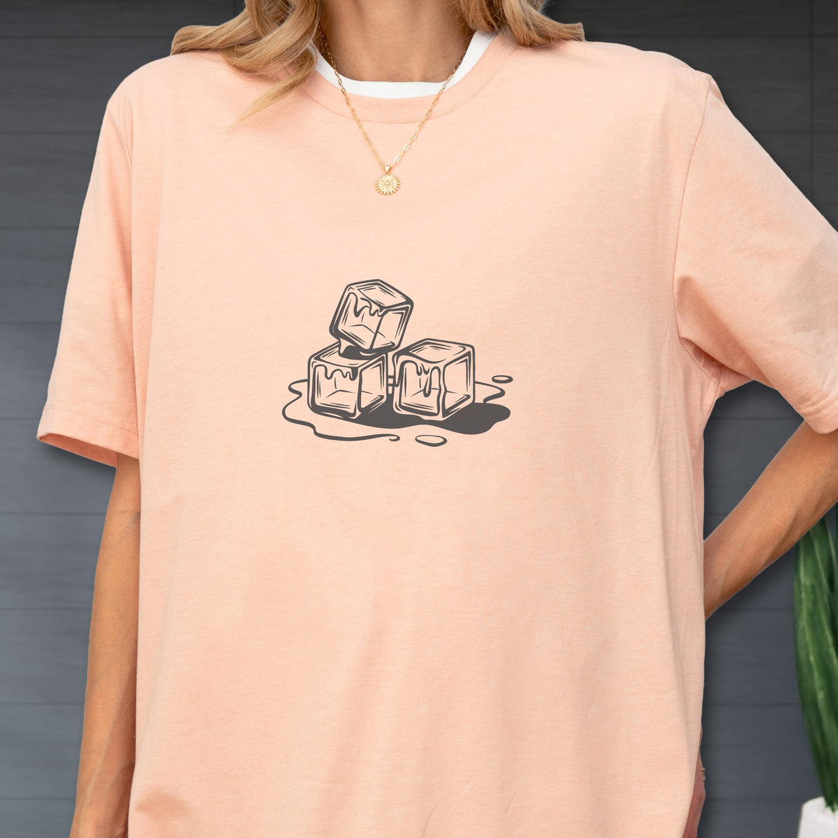 May include: A light peach-colored t-shirt with a white collar. The shirt features a graphic of three melting ice cubes in a black and white illustration. The ice cubes are stacked and surrounded by water droplets.