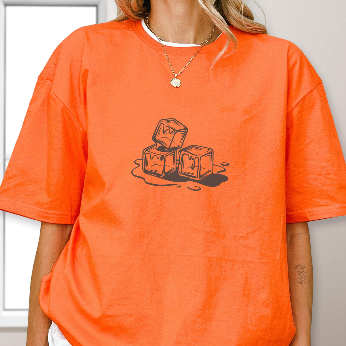 May include: Orange t-shirt with a white crew neck collar. The shirt features a graphic of three melting ice cubes in a brown outline. The ice cubes are dripping onto a puddle.