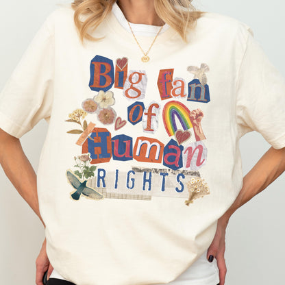 May include: Cream-colored t-shirt with the text "Big fan of Human Rights" in a collage style. The design includes a rainbow, hearts, and floral elements. The shirt has short sleeves and a crew neck.
