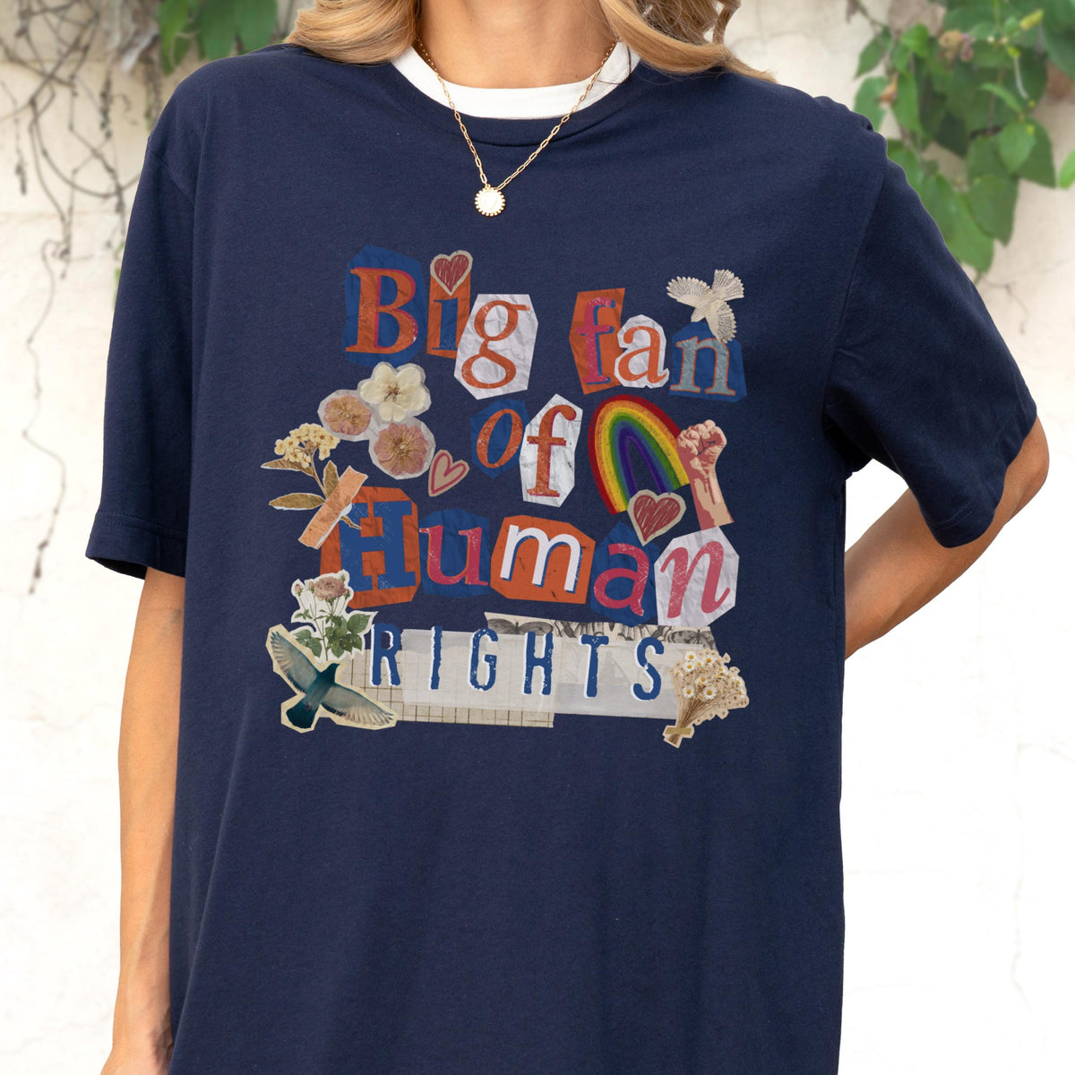May include: Navy blue t-shirt with a white collar featuring the text "Big fan of Human Rights" in a collage style. The design includes a rainbow, hearts, and floral elements. A gold necklace is visible.