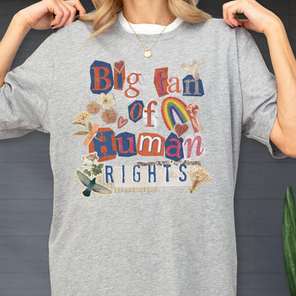 May include: A gray crew neck t-shirt with the text "Big fan of Human Rights" in a colorful collage style. The design includes a rainbow, flowers, and hearts. The shirt has a white collar.