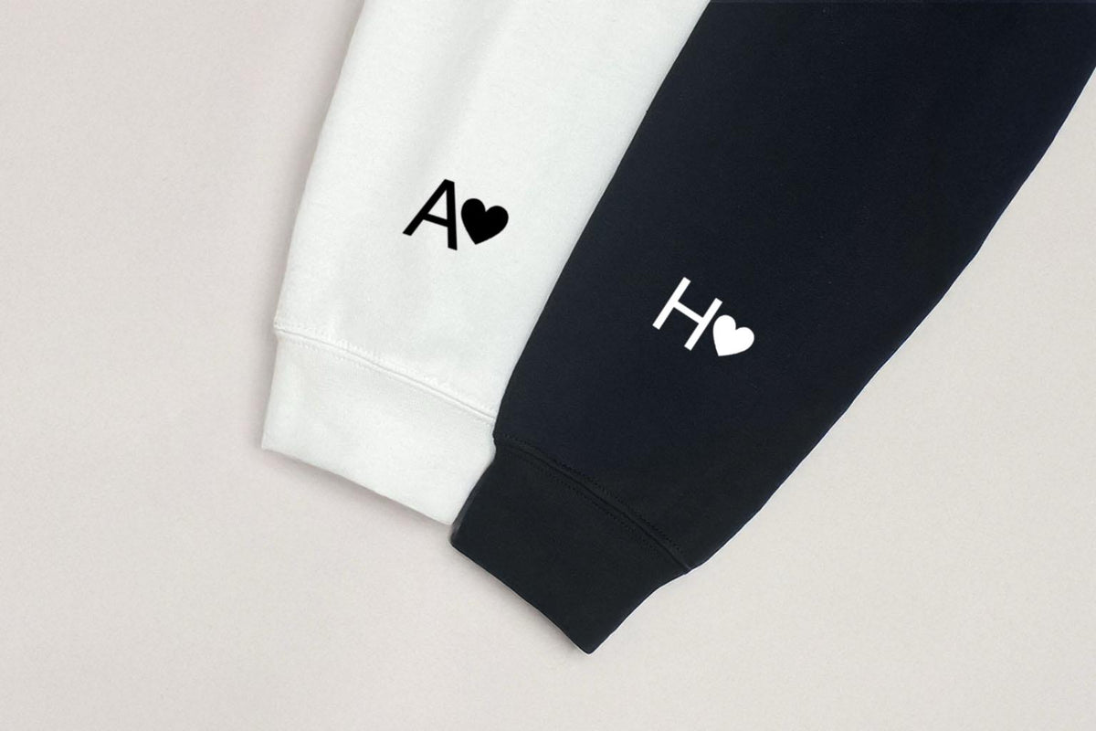 May include: Close-up of two sweatshirt sleeves, one white and one black. The white sleeve has the letter "A" and a heart, while the black sleeve has the letter "H" and a heart. The design is simple and minimalist.