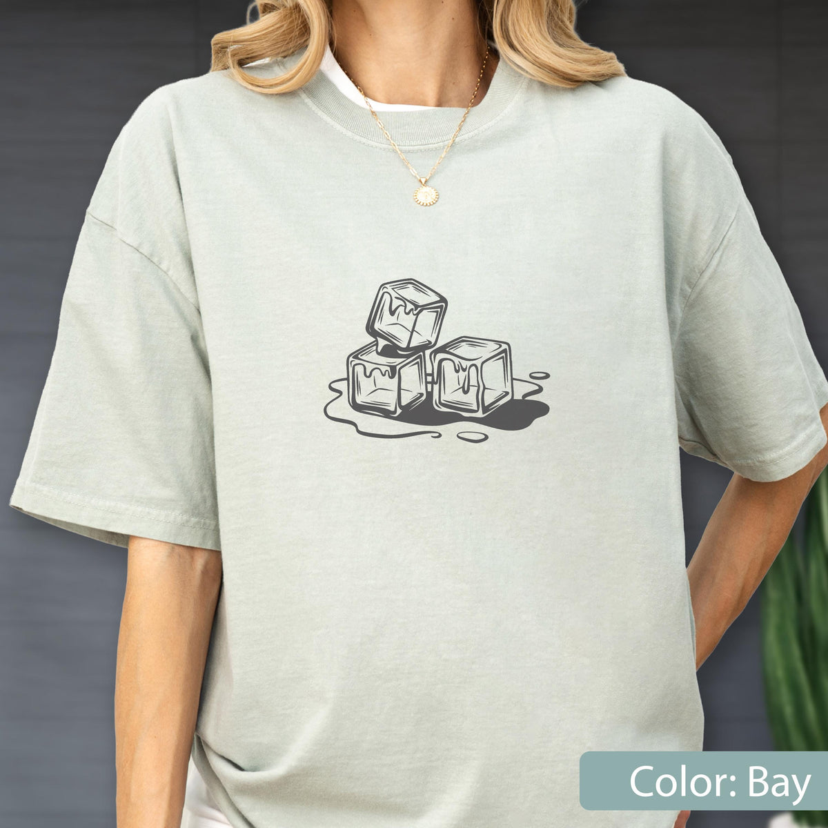 May include: A light green t-shirt with a graphic of three melting ice cubes. The ice cubes are stacked and surrounded by a puddle of water. The shirt is a solid color and has short sleeves.