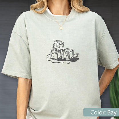 May include: A light green t-shirt with a graphic of three melting ice cubes. The ice cubes are stacked and surrounded by a puddle of water. The shirt is a solid color and has short sleeves.