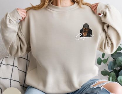 May include: Beige sweatshirt with a graphic of a dog wearing a hat and sunglasses, with the name "Cooper" in cursive. The dog is wearing a black jacket. The sweatshirt has long sleeves and a crew neck.