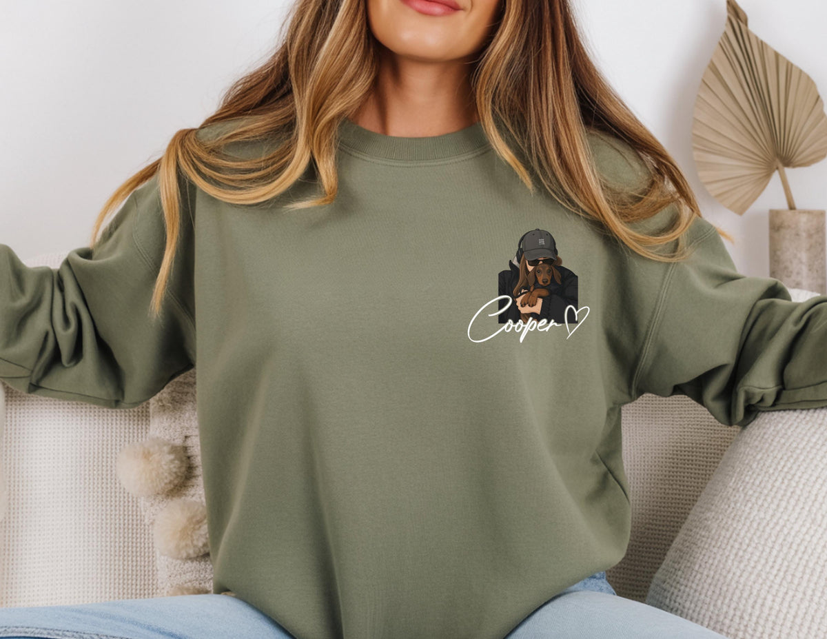 May include: Olive green sweatshirt featuring a graphic of a person holding a dachshund. The graphic includes the name "Cooper" in cursive with a heart. The sweatshirt is a crew neck style.