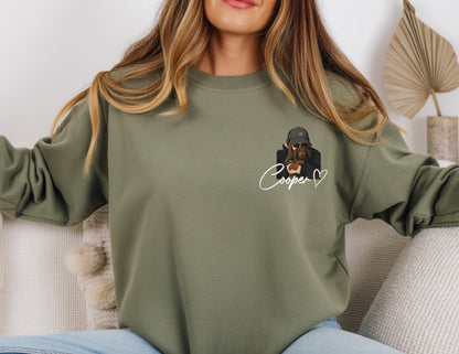 May include: Olive green sweatshirt featuring a graphic of a person holding a dachshund. The graphic includes the name "Cooper" in cursive with a heart. The sweatshirt is a crew neck style.