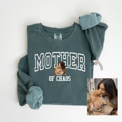 May include: A teal Comfort Colors sweatshirt with the words "MOTHER" and "OF CHAOS" in white block letters. A small image of a woman holding a dog is in the center. The sleeves are cuffed.
