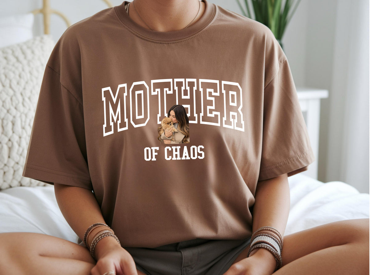 May include: A brown t-shirt with the words "MOTHER OF CHAOS" in white block letters. The shirt features an image of a woman holding a small dog. The t-shirt is a casual wear item.