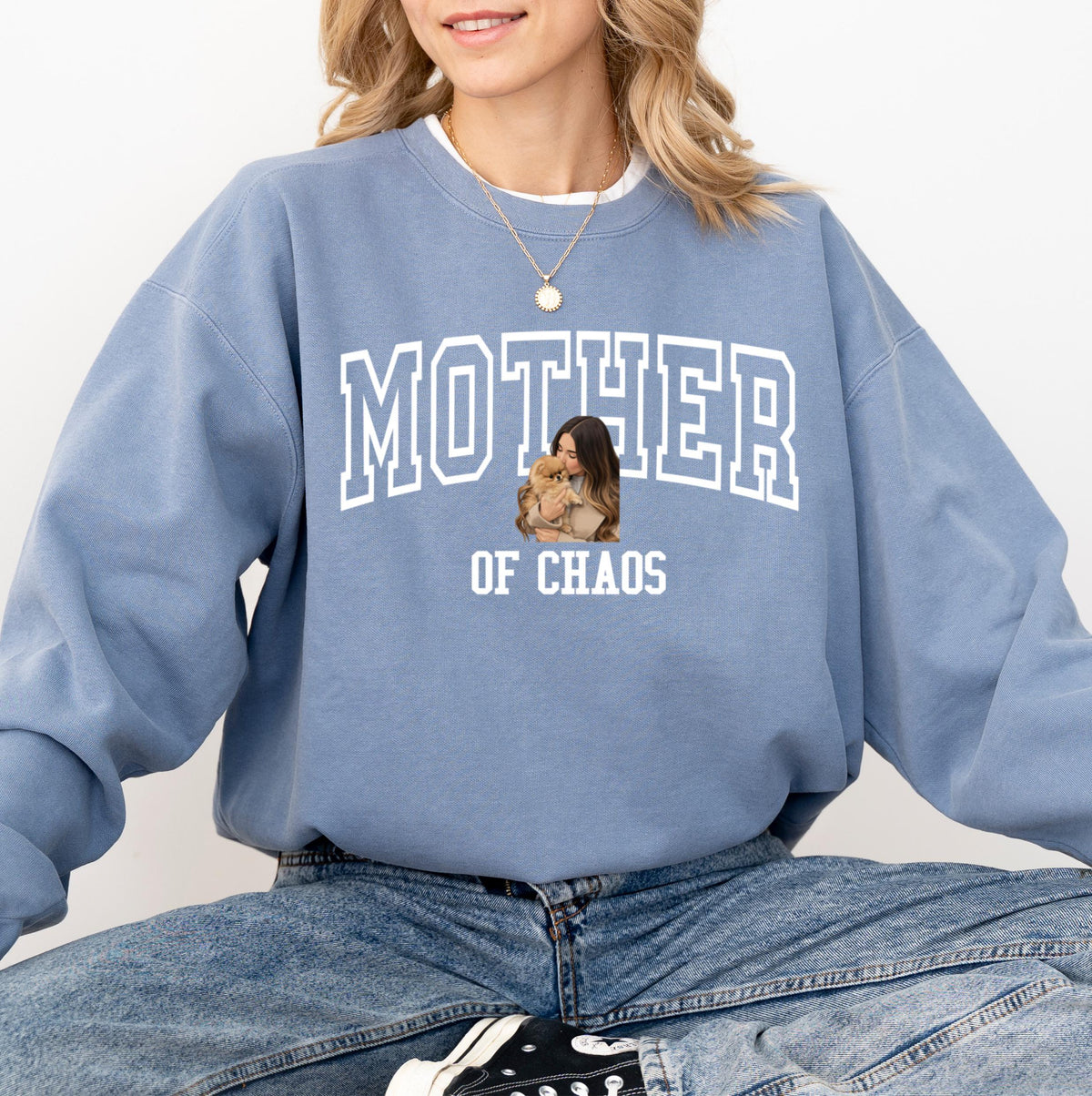 May include: A light blue sweatshirt with the words "MOTHER OF CHAOS" in white block letters. The sweatshirt features a photo of a woman holding a small dog. The model is wearing blue jeans.