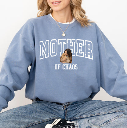 May include: A light blue sweatshirt with the words "MOTHER OF CHAOS" in white block letters. The sweatshirt features a photo of a woman holding a small dog. The model is wearing blue jeans.